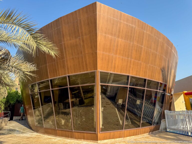 Daytime view of a contemporary Abu Dhabi pavilion with a curved bronze-colored upper shell above dark glazing, framed by palms and low planting.