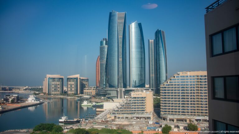 Daytime view of Abu Dhabi’s Etihad Towers and surrounding Corniche skyline seen across marina water and low waterfront buildings.