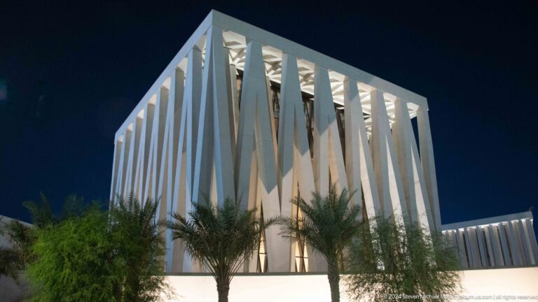 Night exterior of a cubic white Abrahamic Family House worship building on Saadiyat Island, its facade wrapped in deep vertical fins and framed by palm trees.