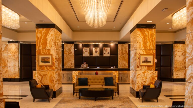 Opulent hotel lobby at Emirates Palace Mandarin Oriental in Abu Dhabi, with polished marble floors and walls, dark wood accents, a large central chandelier, and symmetrical seating flanked by framed portraits.