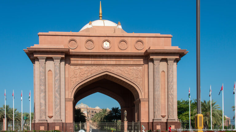 Daytime frontal view of the large ceremonial Emirates Palace entrance gate in Abu Dhabi, with a pointed central arch and flags lining the gateway.