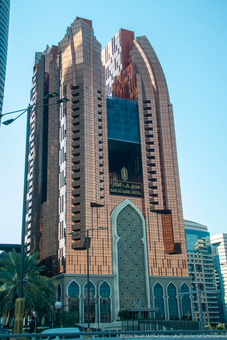 Telephoto view of a modern high-rise in Abu Dhabi with a large turquoise pointed-arch motif and contrasting dark and light vertical facade stripes.