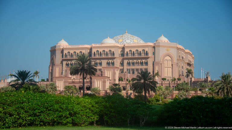 Emirates Palace Mandarin Oriental in Abu Dhabi seen by day behind palm trees and landscaped gardens, with its long facade and central dome visible.