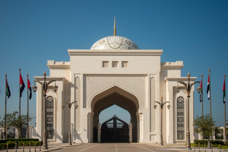 Frontal daytime view of the white Wahat Al Karama entrance gateway in Abu Dhabi, with a central pointed arch and geometric detailing, approached along a roadway lined with UAE flags on both sides.