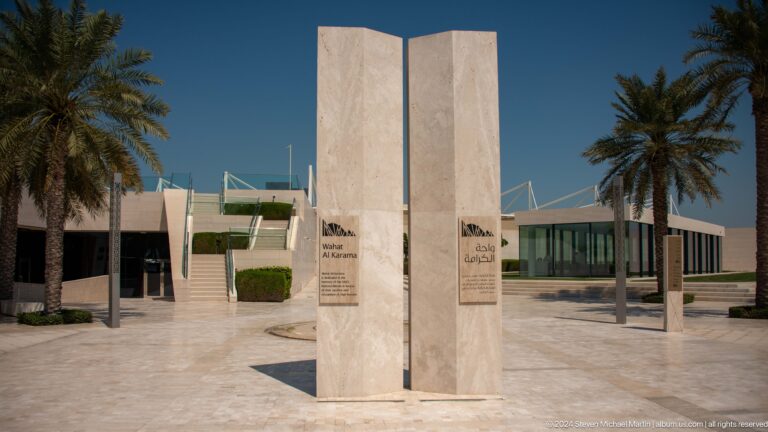 Minimalist vertical stone monument of two pale slabs standing in an open paved plaza at Wahat Al Karama, with low modern buildings and palm trees in the background.