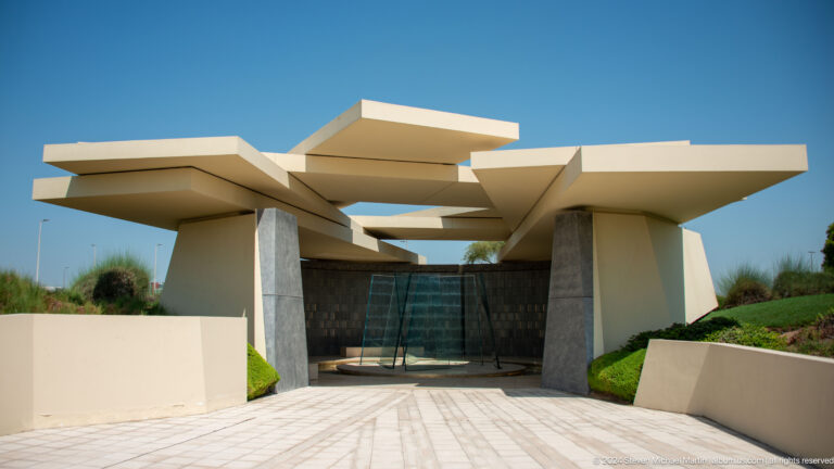 Front view of the Wahat Al Karama visitor pavilion in Abu Dhabi, showing a low modern building with sharply angled roof planes projecting over recessed dark glass doors.