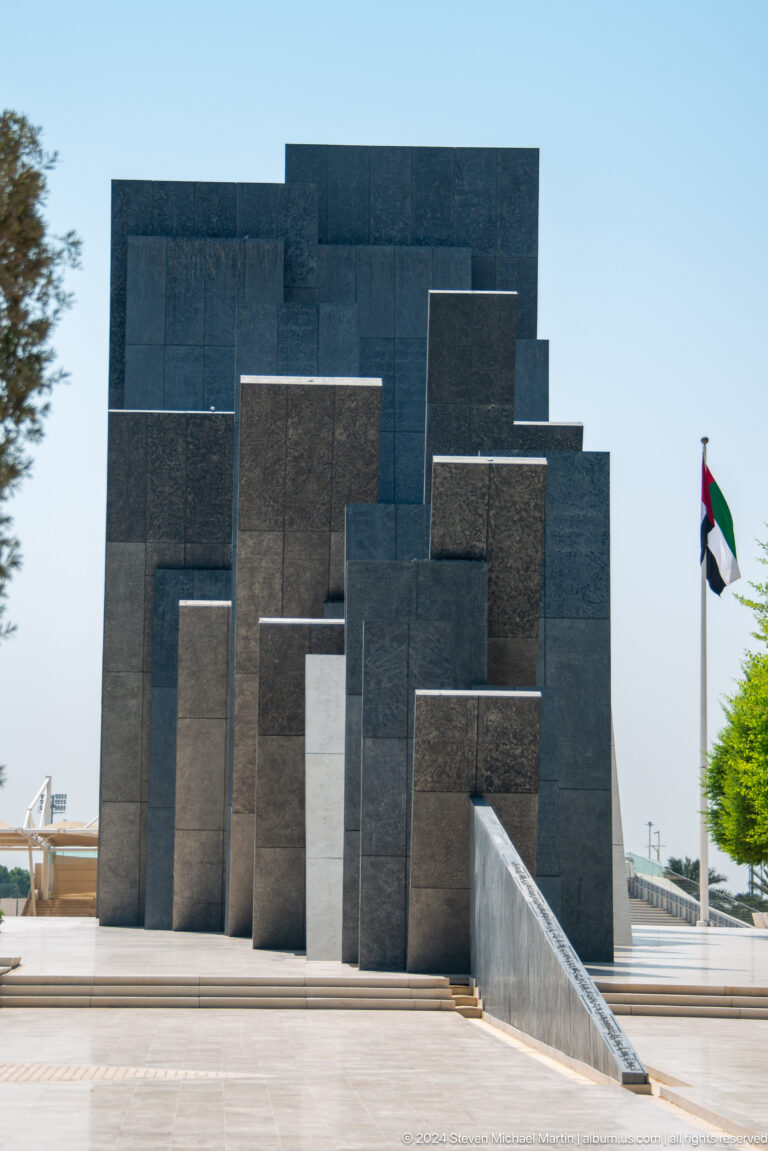 Close view of tall, dark memorial tablets at Wahat Al Karama in the UAE, leaning at varied angles with a UAE flag visible at the edge of the scene.