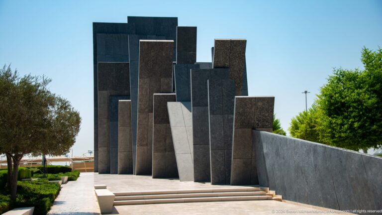 Wide view of Wahat Al Karama in Abu Dhabi, showing a sloped stone walkway leading toward a dense group of overlapping, leaning dark slabs.