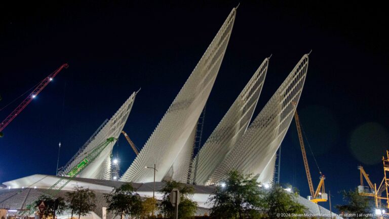 Low-angle nighttime view of the Abrahamic Family House complex on Saadiyat Island, showing three pale triangular roof forms rising against a black sky with construction cranes visible behind.