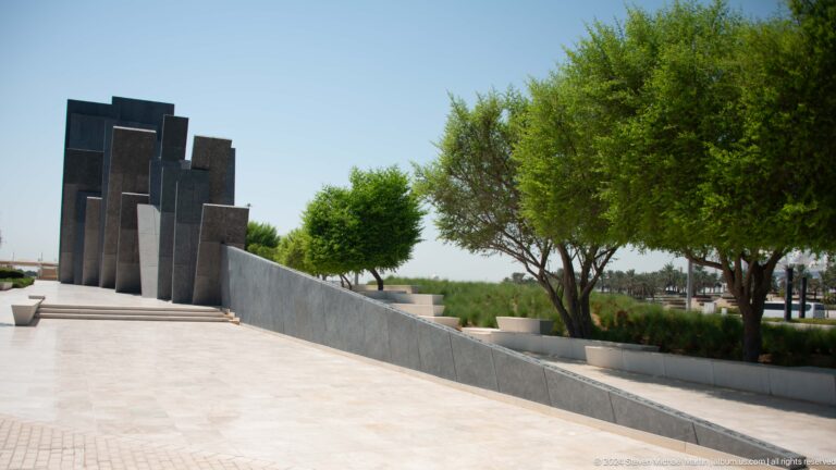 Long pale walkway beside the Wahat Al Karama memorial installation, receding toward dark tablets and lined with evenly spaced green trees.
