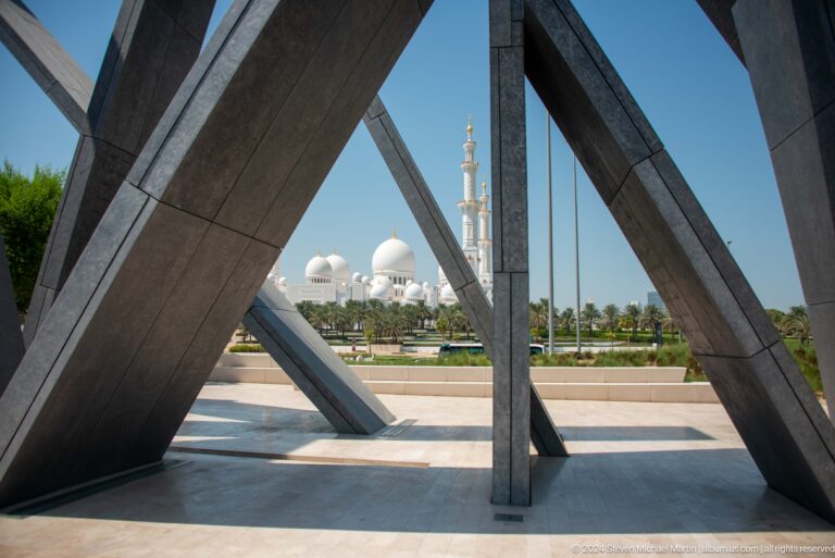 Low view between the dark leaning slabs of Wahat Al Karama framing the white domes and a minaret of Sheikh Zayed Grand Mosque in the distance.