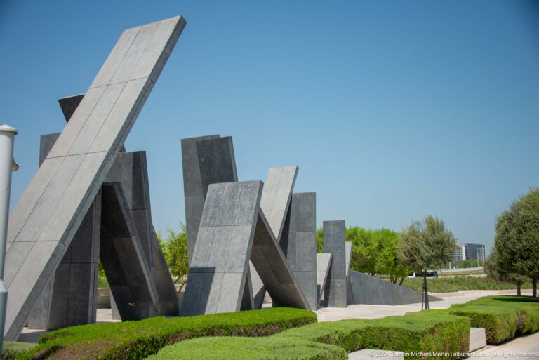 Oblique daytime view of Wahat Al Karama in Abu Dhabi, with a long row of leaning dark slabs stretching across a green lawn bordered by trees and low planting.