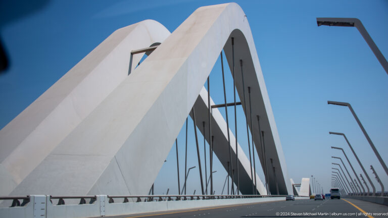 Tight view of Sheikh Zayed Bridge showing a broad white arch curving across a blue sky with vertical black cables descending to the roadway deck.