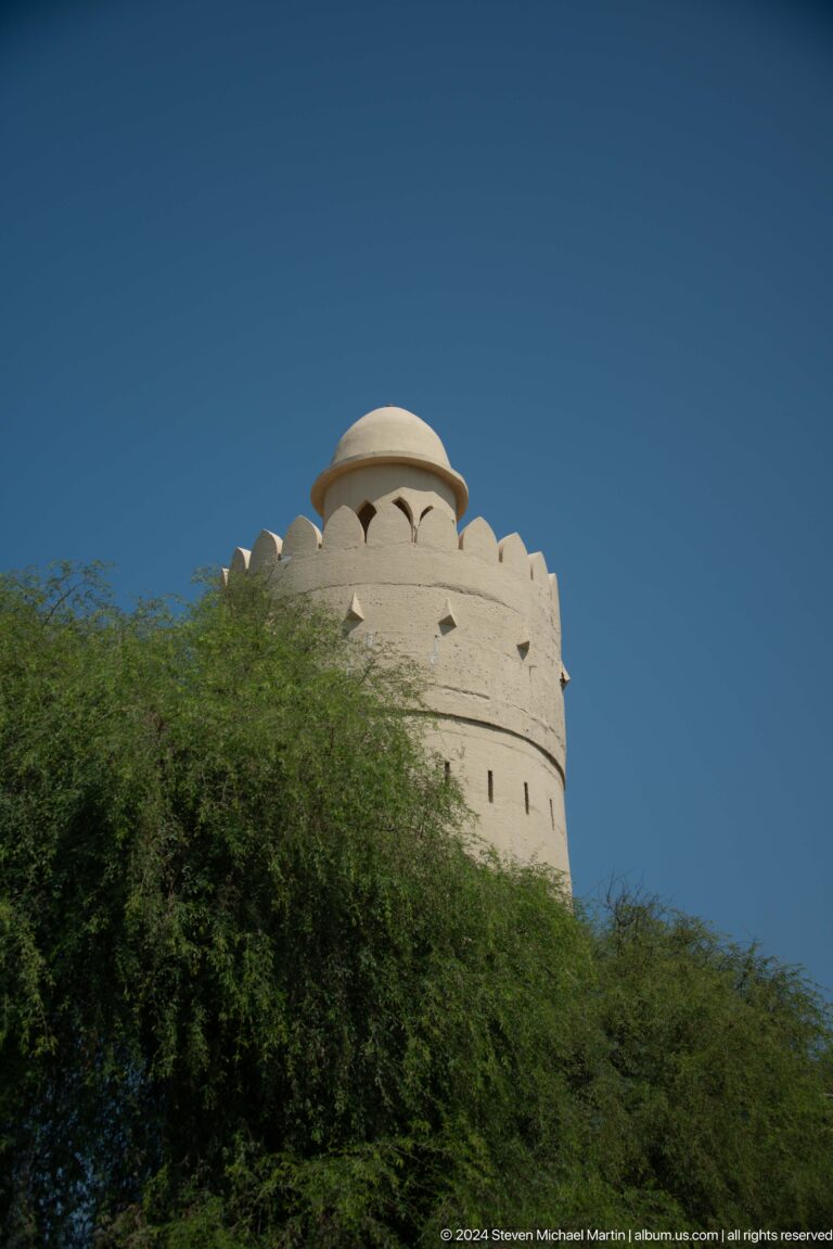 Daytime view of the pale cylindrical Al Maqta Watchtower in Abu Dhabi, with thick walls, narrow slit openings, and a crenellated parapet partly screened by trees.