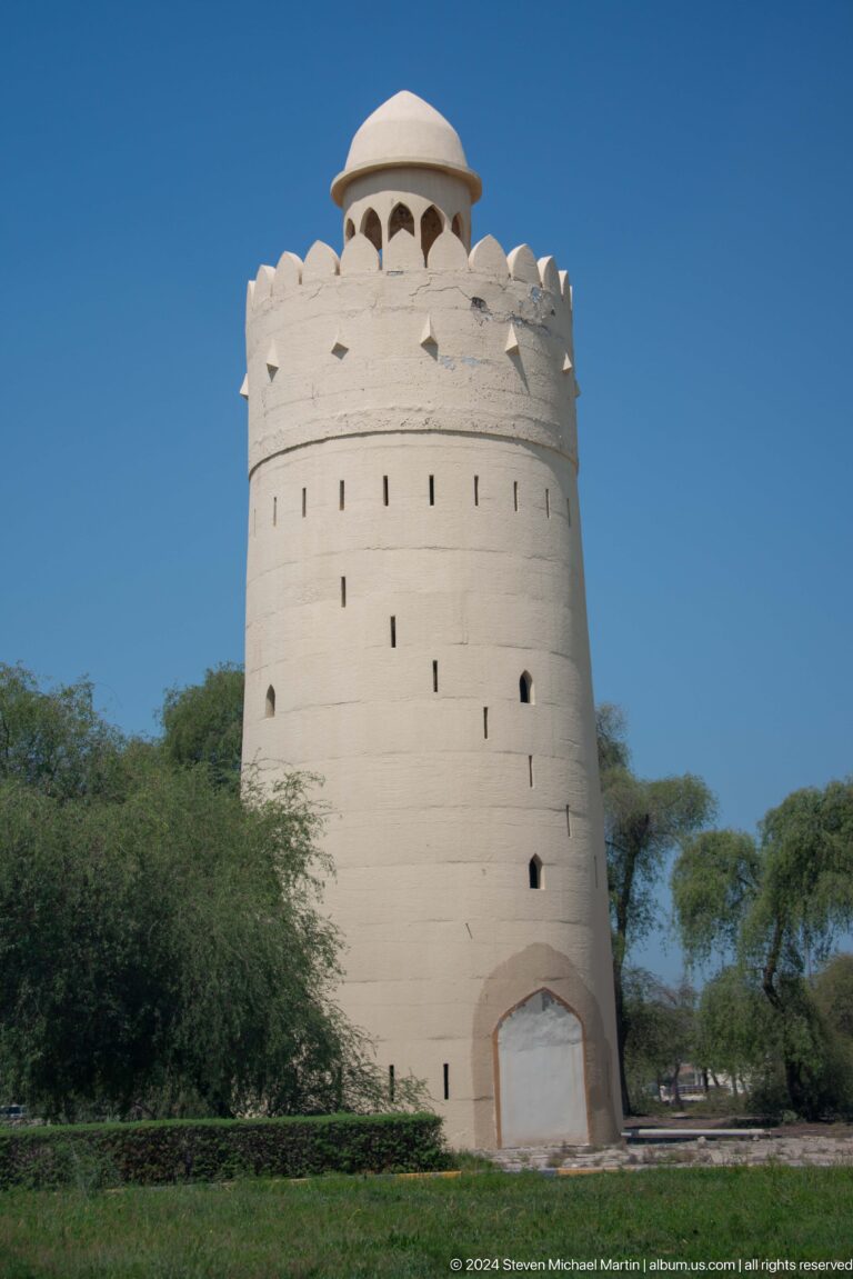 Close view of the upper section of Al Maqta Watchtower in Abu Dhabi, showing narrow vertical loopholes in thick pale walls above surrounding green trees.