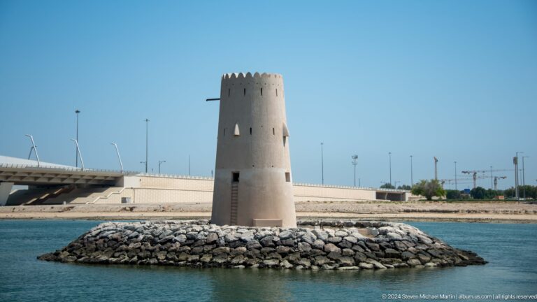 Pale cylindrical watchtower standing alone on a low rock-lined island surrounded by calm blue water at the historic Maqta crossing near Abu Dhabi.