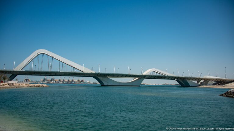 Side view of Sheikh Zayed Bridge’s white wave-like arches spanning the blue Maqta Channel near historic Maqta in Abu Dhabi.