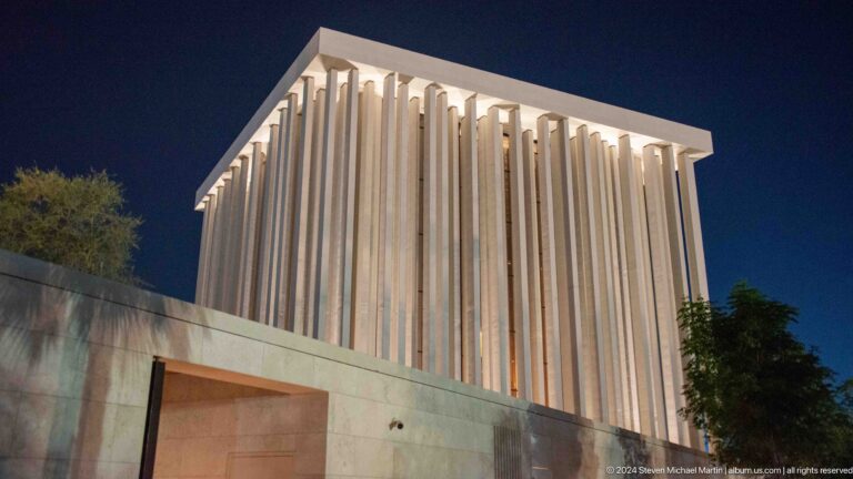 Night view of a luminous cubic worship building at the Abrahamic Family House in Abu Dhabi, with warm light shining through tall vertical facade ribs above a solid base.
