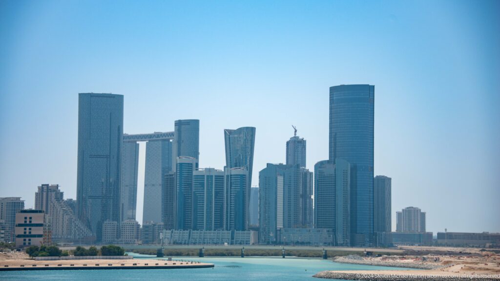 Wide view of Abu Dhabi’s modern waterfront skyline with a continuous band of rectangular high-rise towers seen across calm water.