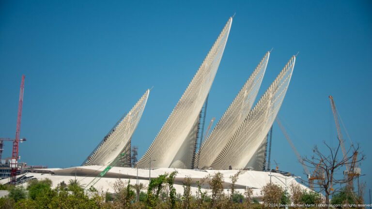 Daytime view of three asymmetrical, perforated triangular forms rising above a low building at the Wahat Al Karama memorial precinct in Abu Dhabi.