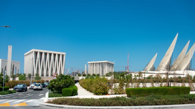 Wide daytime view of Wahat Al Karama in Abu Dhabi, with white finned buildings on the left, tall tapering sculptural forms on the right, and landscaped paths with low planting in the foreground.