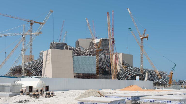 Daytime view of the Guggenheim Abu Dhabi under construction on Saadiyat Island, with multiple cranes surrounding large unfinished concrete and steel forms.