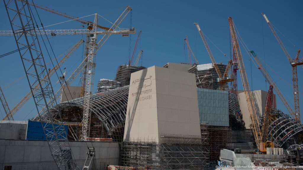 Telephoto view of the Guggenheim Abu Dhabi under construction, with unfinished steel and concrete masses behind perimeter walls and multiple crane booms overlapping the skyline.