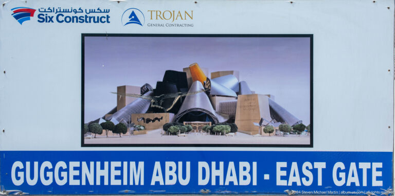 Close-up of a Guggenheim Abu Dhabi construction billboard showing an architectural rendering of clustered sculptural museum volumes with the text “GUGGENHEIM ABU DHABI” on a blue background.