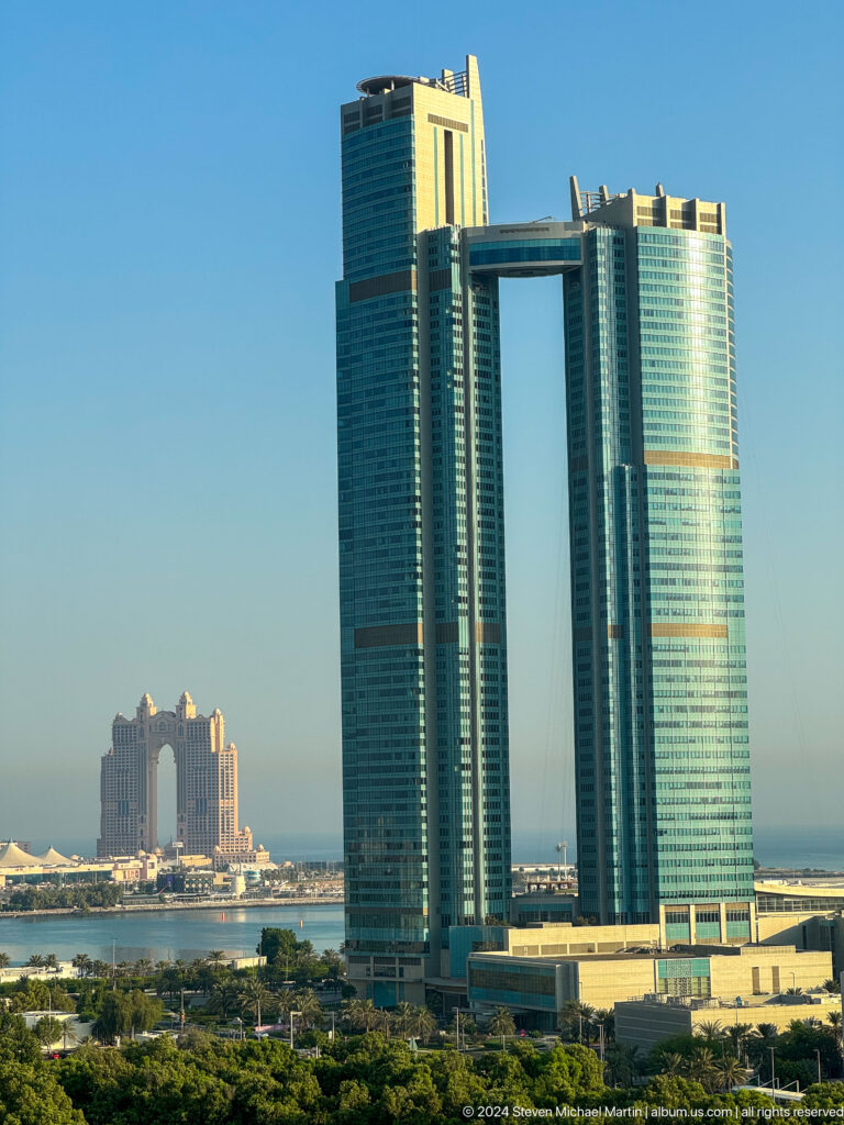 Telephoto view of Abu Dhabi skyline showing the reflective curved Etihad Towers beside the ornate Fairmont Marina Abu Dhabi with its central arch on the waterfront.