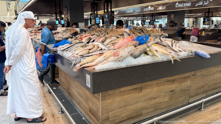 Wide interior view of fresh seafood stalls at Souq Al Mina Fish Market in Abu Dhabi, with many whole fish on crushed ice and vendors serving customers behind the counter.