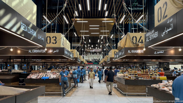 Central aisle of Souq Al Mina fish market with customers walking between long fish counters, beneath large numbered bilingual signs and bright linear ceiling lights.