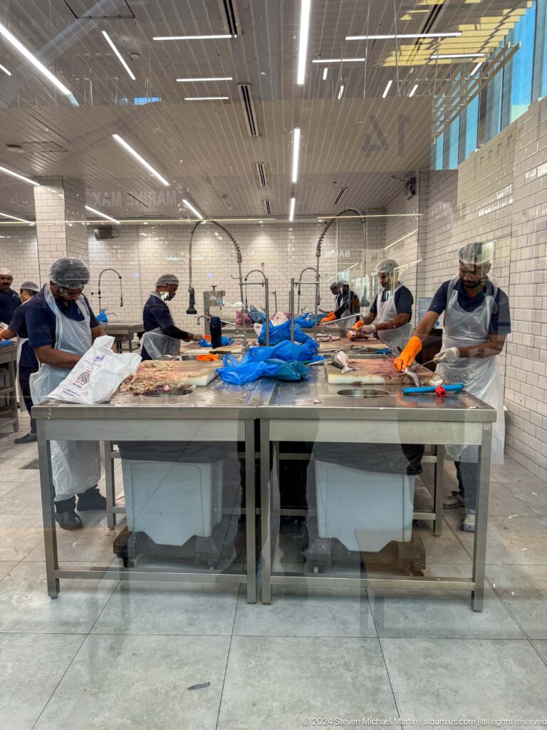 Workers wearing aprons, gloves, and hair coverings clean fish on large stainless-steel tables in a tiled preparation area at Souq Al Mina.