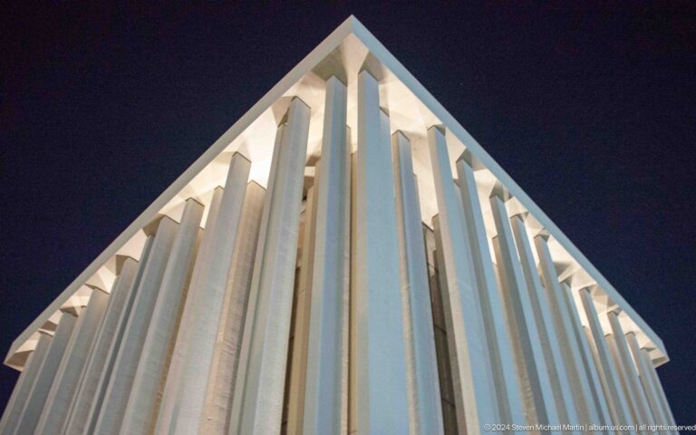 Low-angle view up the sharp corner of an Abrahamic Family House worship building, showing deep white fins projecting from the facade and casting strong bands of light and shadow against the dark sky.