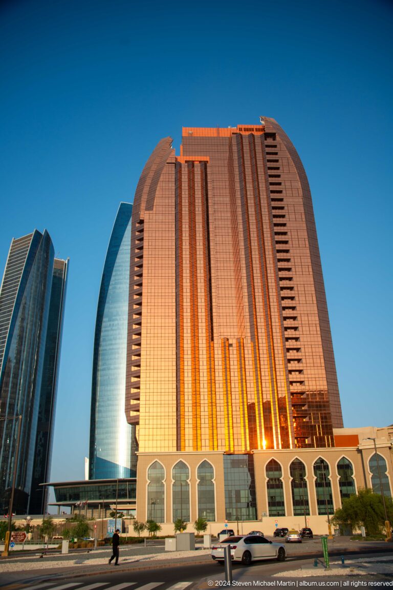 Telephoto view of a tall copper-gold Abu Dhabi high-rise at sunset, its reflective facade glowing in warm light with a narrow central recess emphasizing vertical height.