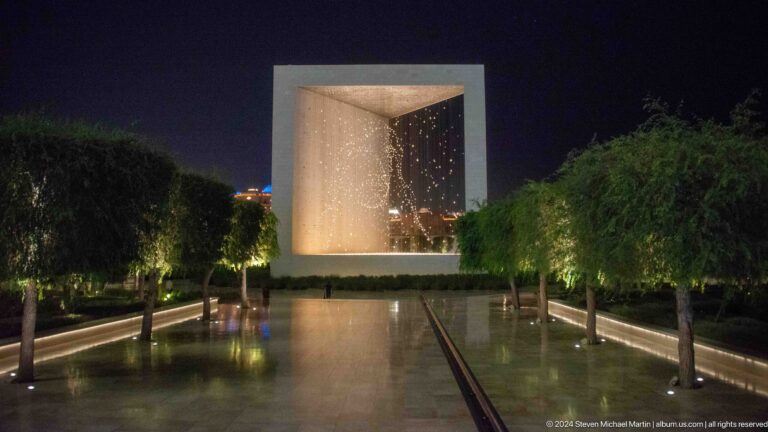 Night view of The Constellation at The Founder’s Memorial in Abu Dhabi, a large open cube filled with suspended lights reflecting in a calm water pool.