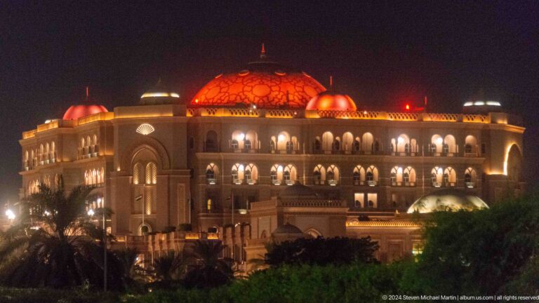 Night view of Emirates Palace Mandarin Oriental in Abu Dhabi, with a warm red-orange central dome and glowing arcades extending across the facade.