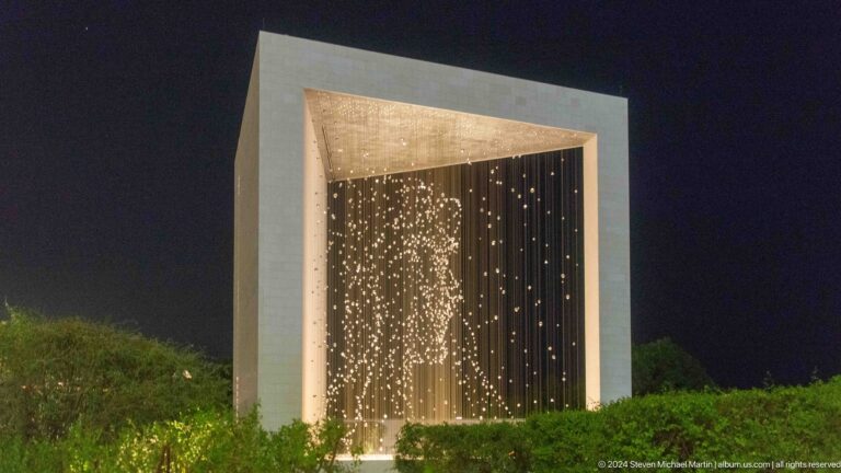 Night view of The Constellation at The Founder's Memorial in Abu Dhabi, a warm field of suspended lights contained within a large pale rectangular frame above a low hedge against a dark sky.