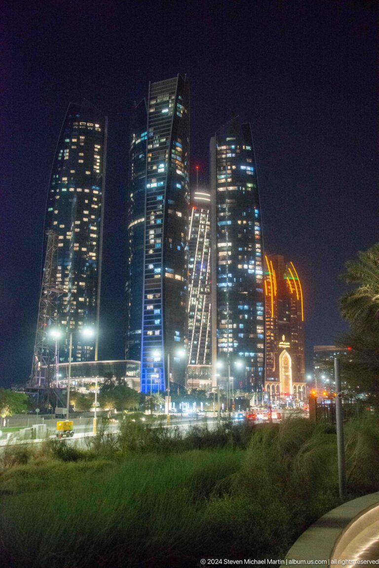 Street-level view of Abu Dhabi high-rises at night with patterned white facade lights, moving traffic, and an orange-lit vertical feature on the right.