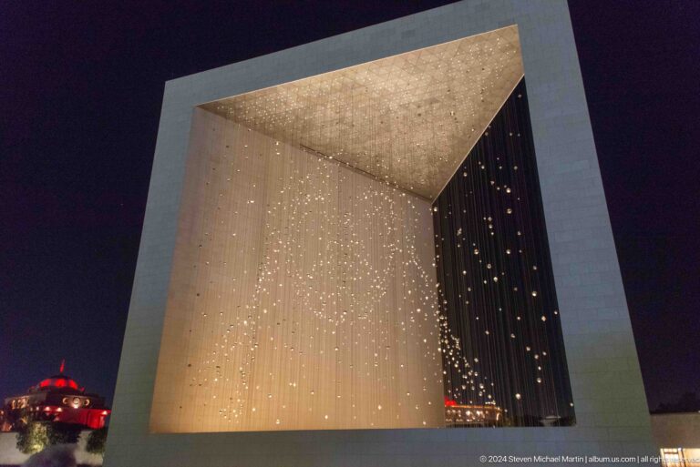 Low-angle view up through the open corner of The Constellation at The Founder's Memorial, showing dense suspended lights forming a three-dimensional portrait within a white structural frame at night.