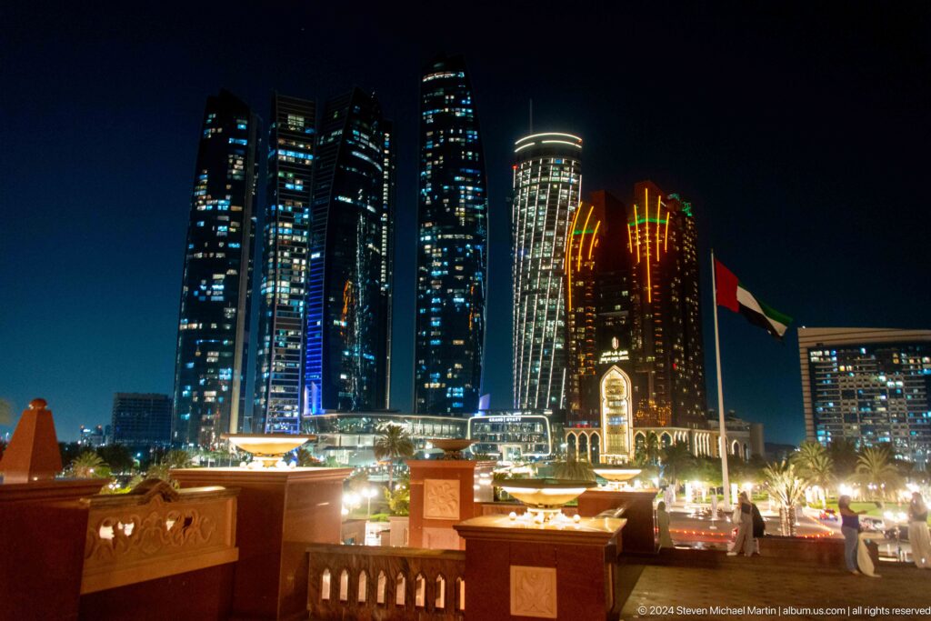 Night view of Abu Dhabi with brightly lit modern high-rises behind a low white complex flying UAE flags.