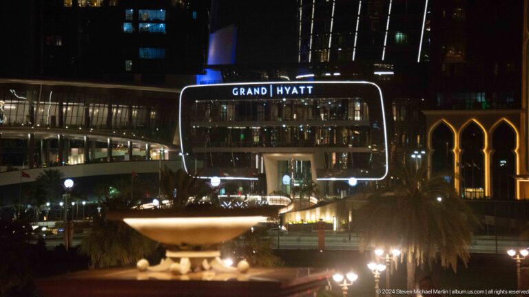 Night view of the Grand Hyatt Abu Dhabi entrance on the Corniche, with the illuminated GRAND HYATT sign set into a curved, brightly lit canopy amid surrounding towers.