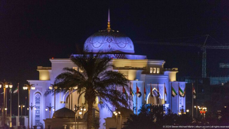 Night view of the Qasr Al Watan entrance gateway in Abu Dhabi, a domed, pointed-arch structure lit in blue-violet with a palm tree silhouetted against the illuminated facade.