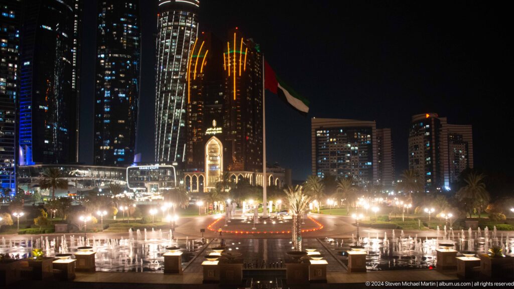 Wide night view of a brightly lit Abu Dhabi waterfront plaza with dense white lights, a tall UAE flag, and modern towers in the background.