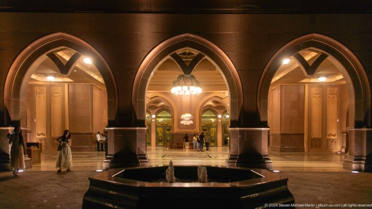 Night view of the Emirates Palace Mandarin Oriental entrance court with a tiered fountain centered in front of three large, warmly lit arches.