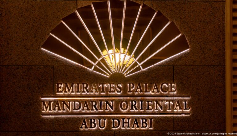 Night view of the Emirates Palace Mandarin Oriental Abu Dhabi sign, with gold illuminated lettering and a sunburst-style emblem above.