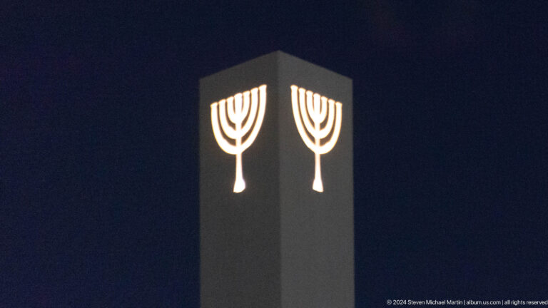 Night view of two illuminated seven-branched menorah emblems mounted near the top of a tall dark rectangular sign.