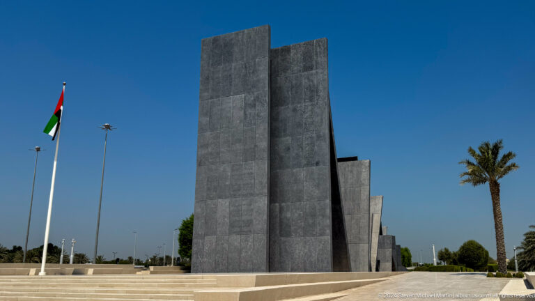 Daytime view of the leaning dark slabs of Wahat Al Karama – The Memorial in Abu Dhabi, with UAE flags at the edge of the paved plaza against a clear blue sky.