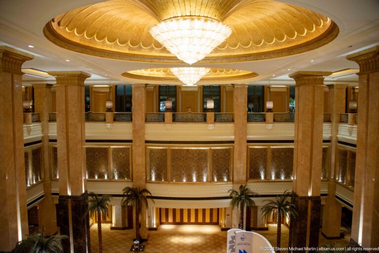 Interior view of the multi-level circular atrium at Emirates Palace with curved balconies, gold-toned decoration, and a large chandelier hanging beneath an ornate dome ceiling.