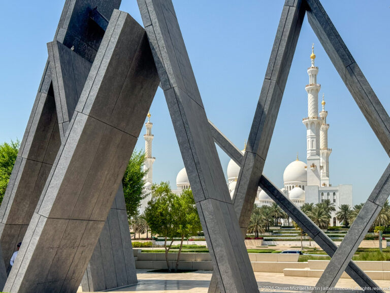 Dark, angled memorial slabs at Wahat Al Karama in Abu Dhabi framing a distant white minaret of Sheikh Zayed Grand Mosque beyond the trees.
