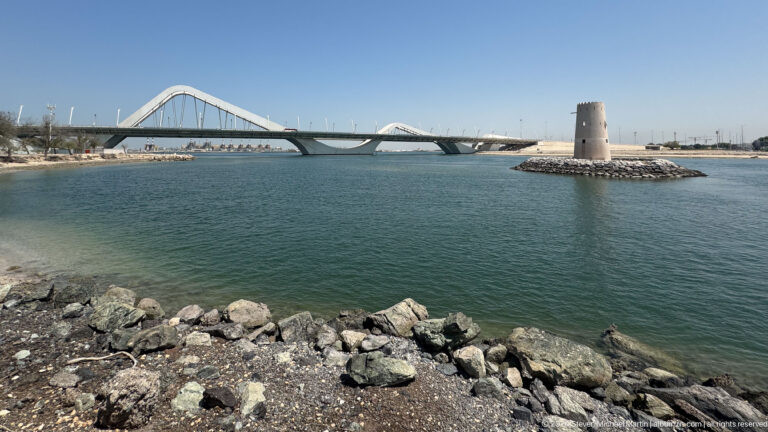 Daytime view over the Maqta Channel toward the pale cylindrical Al Maqta Watchtower on a rocky islet with the white arches of Sheikh Zayed Bridge rising behind it in Abu Dhabi.